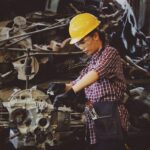 Woman in a yellow hard hat and safety glasses working on a large industrial engine part at a workshop bench