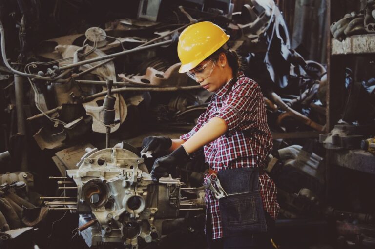 Woman in a yellow hard hat and safety glasses working on a large industrial engine part at a workshop bench