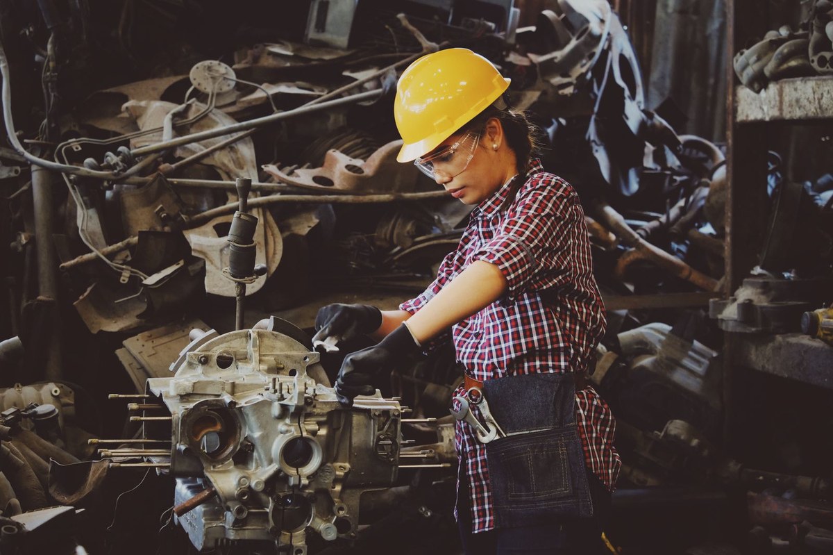 Woman in a yellow hard hat and safety glasses working on a large industrial engine part at a workshop bench